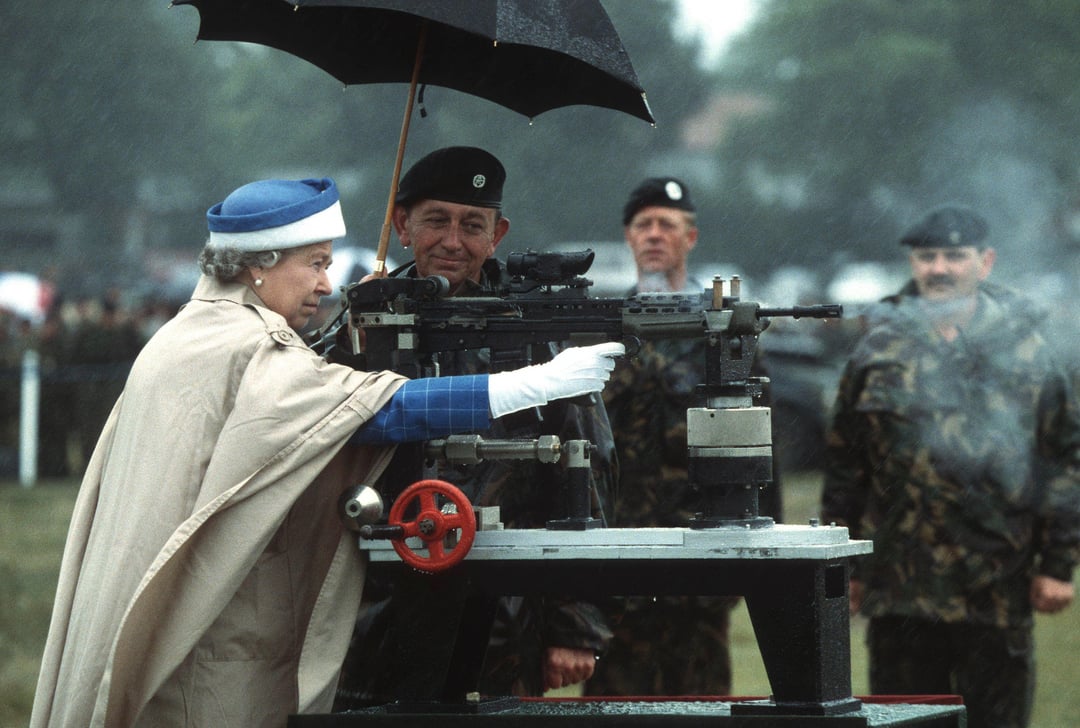 Queen Elizabeth II taking aim and firing a weapon at an Army shooting event, with uniformed soldiers attending under umbrellas in the rain.